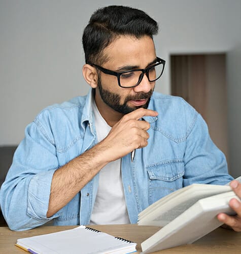 Young indian latin eastern male student in glasses reading book writing notes in textbook at home workplace. High school, university, college exams preparation. Remote distant education concept.