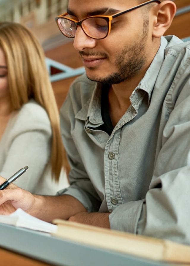 Multi-ethnic group of students sitting at desk in lecture hall of modern college and writing, focus on young Middle-Eastern man wearing glasses, copy space
